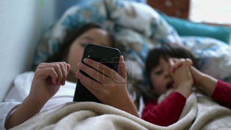 Foto de Primer plano de la mano de la niña que sostiene el dispositivo de teléfono celular en la cama debajo de las sábanas con el hermano pequeño mirando la pantalla junto a ella. dos niños comprometidos con la tecnología moderna - Imagen libre de derechos