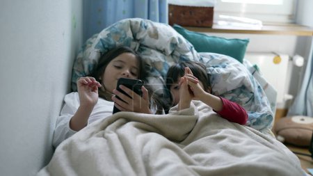 Téléchargez les photos : Enfants partageant écran de téléphone portable dans le lit sous les draps. Petit frère et sœur utilisant un dispositif de technologie moderne - en image libre de droit