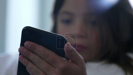 Close-up face of little girl holding and using phone with solemn bored expression. Child watching content online
