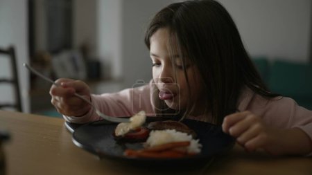 Téléchargez les photos : Petite fille mangeant le déjeuner, enfant de 8 ans appréciant la nourriture à l'heure des repas. Assiette de viande, légumes et riz - en image libre de droit