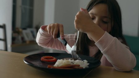 Téléchargez les photos : Petite fille appréciant le dîner fait maison à la maison, dîner enfant de 8 ans, couper la nourriture avec une fourchette et un couteau par elle-même - en image libre de droit
