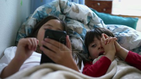 Photo for Close-up little girl's hand holding cellphone device in bed under bedsheets with small brother sibling watching screen next to her. two children engaged with modern technology - Royalty Free Image