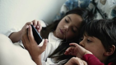 Téléchargez les photos : Frère et sœur Engrossis dans l'écran du téléphone intelligent alors qu'ils sont couchés au lit, frères et sœurs collant sur un appareil numérique - en image libre de droit