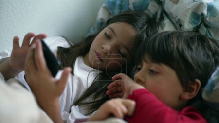 Photo for Young Siblings Sharing Cellphone Screen in Bed, Sister and Brother Looking at Smartphone Together - Royalty Free Image