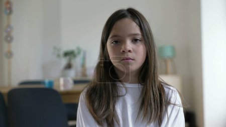 Téléchargez les photos : Portrait d'une petite fille solennelle regardant la caméra avec une expression sérieuse à la maison à l'intérieur, salon, enfant de 8 ans - en image libre de droit