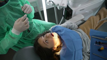 Photo for Apprehensive little girl seated at Dentist office before teeth procedure. Doctor getting ready while 8 year old child observes with anxious preoccupation, overhead view - Royalty Free Image