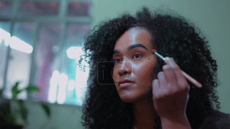 Eine junge schwarze Brasilianerin schminkt sich vor dem Spiegel, eine Person afrikanischer Herkunft mit lockigem Haar, die sich auf das Ausgehen vorbereitet, Gesicht in Großaufnahme