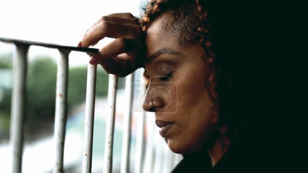 Photo for One depressed black woman feeling sorrow and despair with eyes closed leaning on metal bar on balcony, close-up profile face of desperate middle-aged person feeling sad - Royalty Free Image
