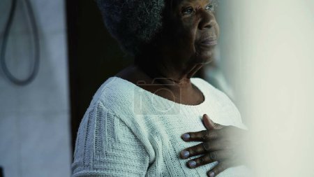 Téléchargez les photos : Candid réfléchie femme afro-américaine senior mettre la main sur la poitrine tout en contemplation mentale profonde regardant la fenêtre vue de la résidence, personne âgée des années 80 - en image libre de droit