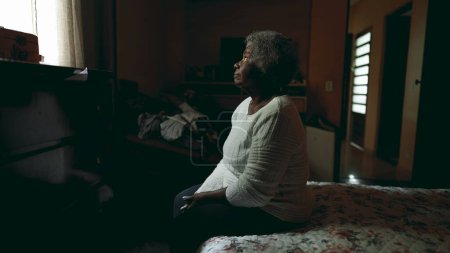 Téléchargez les photos : Lonely dame âgée sud-américaine assis à côté du lit avec la paix regard tranquille à la fenêtre à l'intérieur chambre humble faiblement éclairée contemplant la vie et les souvenirs passés. personne âgée des années 80 - en image libre de droit