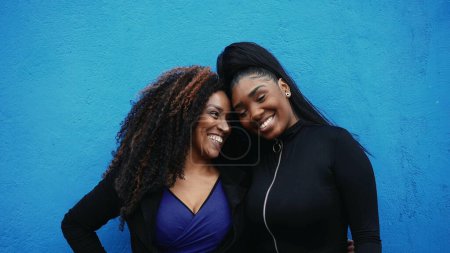 Téléchargez les photos : Bonne mère afro-américaine et adolescente posant ensemble dans l'étreinte de liaison, authentique amour authentique et l'affection entre le parent et la progéniture devant le mur bleu - en image libre de droit