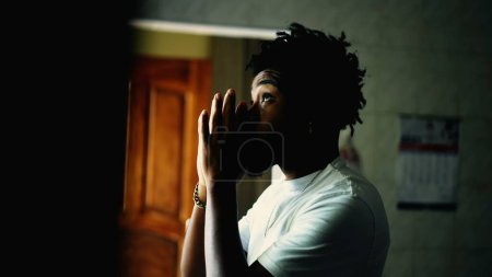 Téléchargez les photos : Jeune homme afro-américain fidèle priant DIEU à la maison par la fenêtre. Un homme noir plein d'espoir dans la PRIÈRE pendant les moments difficiles - en image libre de droit