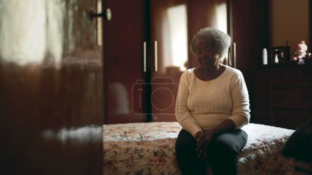 Téléchargez les photos : Candid Senior dame noire sud-américaine avec les cheveux gris assis au chevet avec regard contemplatif réfléchissant à la vieillesse et les défis de la vie, humble fond de maison - en image libre de droit