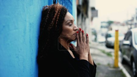 Photo for One anxious middle-aged woman Praying to GOD standing outside leaning on blue wall in urban street feeling desperate. Spiritual person asking for divine help with HOPE and FAITH - Royalty Free Image
