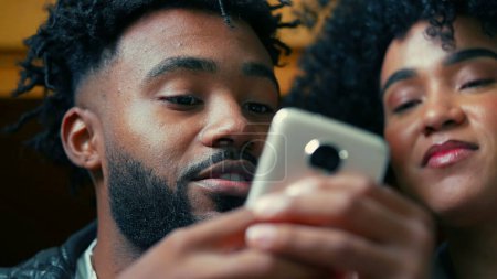 African American young couple close-up faces staring at cellphone device in foreground. People watching entertainment media on phone screen, consuming entertainment media
