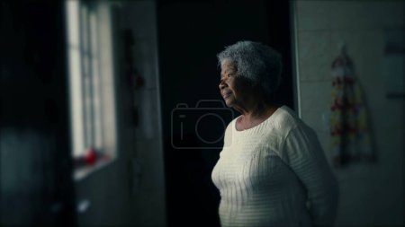Foto de Una anciana contemplativa de 80 años que estaba de pie junto a la ventana en una humilde residencia con expresión reflexiva. Pensativo señora mayor con el pelo gris en la soledad tranquila - Imagen libre de derechos