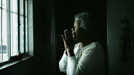 Pieuse Religieusement pieuse femme afro-américaine âgée debout près de la fenêtre de la cuisine dans la PRIÈRE à la recherche d'espoir et de foi. Une dame spirituelle noire aux cheveux gris
