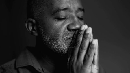Ein ernsthafter älterer schwarzer Mann in tiefem Gebet, der in schwierigen Zeiten um göttliches Eingreifen bittet, Nahaufnahme eines südamerikanischen Seniors afrikanischer Abstammung in ruhiger, kontemplativer Monochromie
