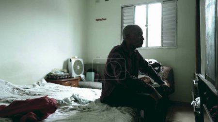 Téléchargez les photos : Un cher homme d'âge mûr assis au chevet d'un lit dans une pièce sombre et lunatique aux prises avec la pauvreté et la dépression, personne réfléchie d'ascendance africaine - en image libre de droit