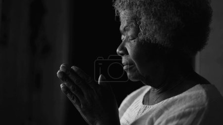 Foto de Una devota religiosa negra anciana ORANDO a DIOS en casa por ventana en monocromático, blanco y negro. Espiritual afroamericana anciana en meditación profunda - Imagen libre de derechos