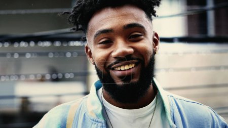Ein glücklicher junger schwarzer Mann steht draußen im Nieselregen und lächelt in die Kamera in der städtischen Umgebung, Gesicht in Großaufnahme