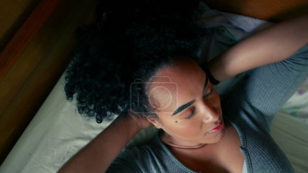 Foto de Una joven negra nerviosa acostada en la cama tratando de relajarse y descansar. Ansiosa persona de 20 años de ascendencia africana abrumada por la vida, sin embargo, se toma un descanso para recuperarse - Imagen libre de derechos