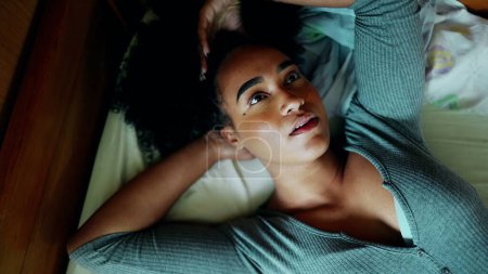 Téléchargez les photos : Une chère jeune latina noire couchée au lit avec une expression contemplative réfléchie regardant la fenêtre et jouant avec les cheveux bouclés réfléchissant à la décision tout en se relaxant à la maison - en image libre de droit