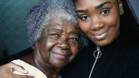 Tender loving moment of African descent granddaughter kissing elderly South American grandmother in the forehead in caring embrace with arm around shoulder, close-up faces