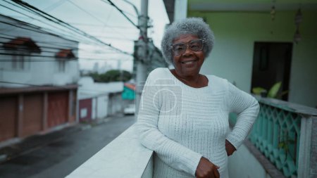 Foto de Una anciana negra feliz de América del Sur de pie en el humilde balcón de la residencia con vistas a la calle urbana en el fondo. Retrato de una persona mayor de 80 años con cabello gris de ascendencia africana - Imagen libre de derechos
