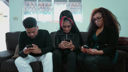 Foto de Tres personas mirando fijamente sus teléfonos sentadas en un sofá en el interior hipnotizadas por la tecnología que representa el aislamiento social y la adicción al teléfono. Afroamericanos enviando mensajes, solos juntos - Imagen libre de derechos