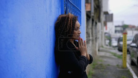 Photo for One Hispanic black latina Woman of African Descent in Mental Contemplation During Challenging Times seeking divine help and support gazing upwards to SKY in PRAYER standing in street outside - Royalty Free Image