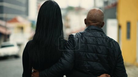 Photo for Tracking shot of a middle-aged father walking with his teenage daughter outside in urban street during drizzle rain. African American mature person bonding with family while walking forward - Royalty Free Image