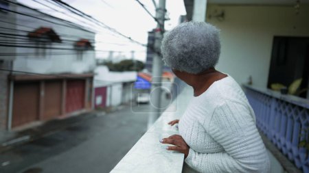 Téléchargez les photos : Une femme sud-américaine âgée observant le quartier depuis le balcon de la résidence. 80 ans personne âgée regardant la rue urbaine d'en haut avec une expression heureuse - en image libre de droit