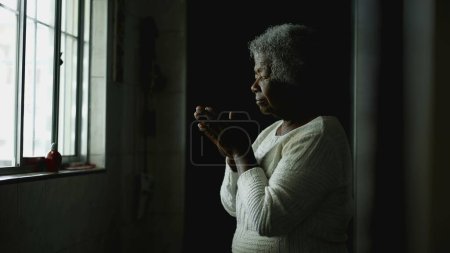 Foto de Anciana Afroamericana Espiritual en Meditación Profunda, escena de oración de esperanza de una anciana negra con el pelo gris de pie junto a la ventana de la cocina con los ojos cerrados - Imagen libre de derechos
