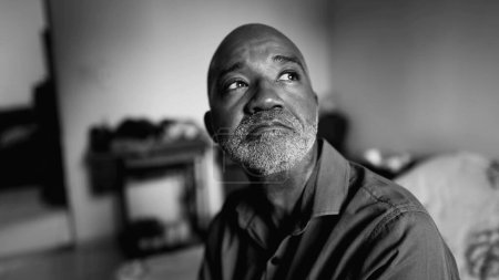 Thoughtful depressed African American senior man with sad emotion struggles in solitude in moody bedroom in dramatic black and white, monochrome portrait
