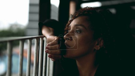 Foto de Una triste y pensativa mujer negra de mediana edad mirando a la vista desde el balcón en profunda contemplación melancólica. Cara de cerca de una mujer afroamericana sintiéndose perdida en el pensamiento, reflexionando sobre los desafíos de la vida - Imagen libre de derechos