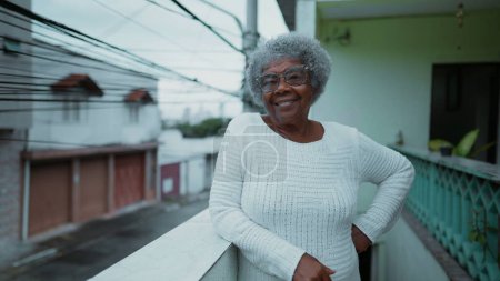 Photo for One happy elderly black woman from South America standing at humble residence balcony overlooking urban street in background. Portrait of 80s senior person with gray hair of African descent - Royalty Free Image