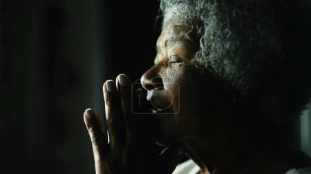 Téléchargez les photos : Religieuse afro-américaine âgée fermant les yeux dans Prière par fenêtre, profil rapproché d'une dame noire âgée aux cheveux gris en méditation profonde - en image libre de droit