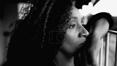 Sad Depressed 50s woman leaning on metal bar at home balcony feeling lost in thought in deeo mental rumination in intense dramatic black and white. Portrait of contemplative South American latina