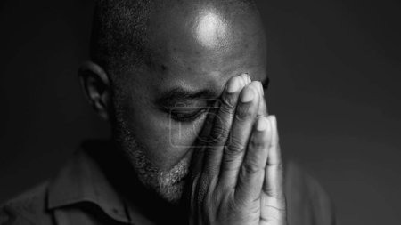 Ein ernsthafter älterer schwarzer Mann in tiefem Gebet, der in schwierigen Zeiten um göttliches Eingreifen bittet, Nahaufnahme eines südamerikanischen Seniors afrikanischer Abstammung in ruhiger, kontemplativer Monochromie