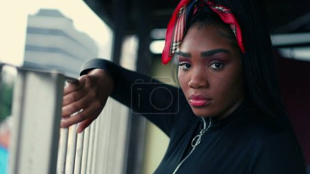 Foto de Retrato de una joven latina negra mirando a la cámara con expresión seria de pie en el balcón de casa apoyado en la barra de metal y mirando a la cámara, persona de 20 años de ascendencia africana - Imagen libre de derechos