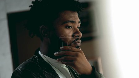 Téléchargez les photos : Jeune homme afro-américain en réflexion mentale debout par la fenêtre avec un regard contemplatif réfléchissant à la solution aux problèmes. Personne dans le processus de remise en question des croyances - en image libre de droit