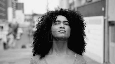 Téléchargez les photos : Une jeune femme noire pleine d'espoir aux cheveux bouclés debout dans la rue regardant le ciel avec espoir et foi en noir et blanc. Afro-Américain sentant la présence de DIEU - en image libre de droit