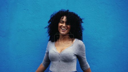 Foto de Un hapyp joven negro brasileña auténtica vida real risa y sonrisa de pie en el fondo azul al aire libre en la calle de la ciudad sonriendo a la cámara, cabello rizado empoderado persona afrodescendiente - Imagen libre de derechos