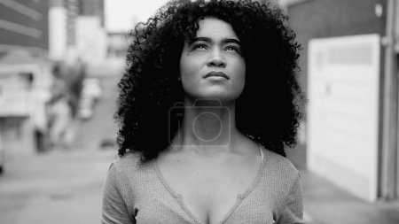 One hopeful young black woman with curly hair standing in street looking up at sky with HOPE and FAITH in black and white. African American person feeling the presence of GOD