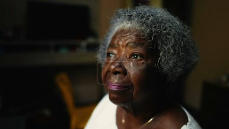 Téléchargez les photos : Gros plan d'une femme afro-américaine âgée regardant la fenêtre à l'intérieur dans la solitude. Une dame noire plus âgée pensif réfléchi avec les cheveux gris en contemplation - en image libre de droit