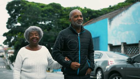 Téléchargez les photos : Fils afro-américain adulte marchant avec une mère âgée en ville, dame âgée dans ses années 80 bras d'armes avec gardien à l'extérieur - en image libre de droit