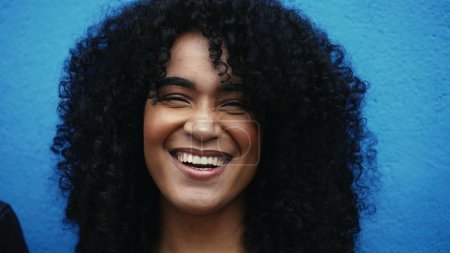 Foto de Una alegre joven latina negra con el pelo rizado sonriendo a la cámara en el fondo azul en el ambiente de la ciudad. primer plano de la persona sudamericana de ascendencia africana - Imagen libre de derechos