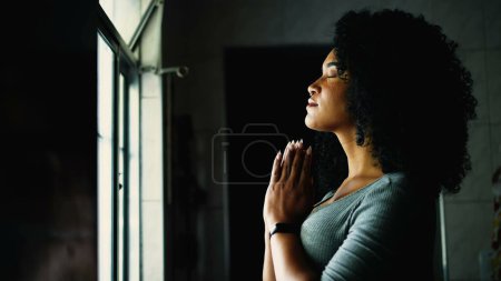 Foto de Una joven negra espiritual de pie junto a la ventana en casa en oración con los ojos cerrados. Afro-americana chica adulta con el pelo rizado comprometido en la meditación profunda, perfil de la cara y las manos apretadas juntos - Imagen libre de derechos