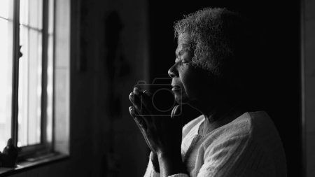 Téléchargez les photos : Dame âgée afro-américaine spirituelle en méditation profonde, noir et blanc Prière à la maison par fenêtre - en image libre de droit